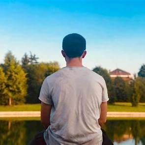 A young person sitting quietly by a calm lakeside at sunset, head bowed and hands clasped, reflecting the loneliness that can follow success.