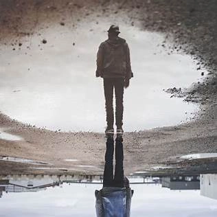 A solitary person standing at the edge of a still puddle, quietly staring at their own reflection — symbolizing the silent introspection and emotional emptiness that can follow personal success.